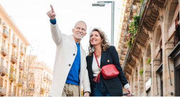 Un couple marche le long d&rsquo;une rue urbaine bord&eacute;e d&rsquo;immeubles historiques. L&rsquo;homme pointe vers le haut et la femme porte un sac &agrave; bandouli&egrave;re rouge.